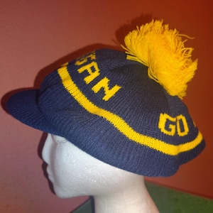 May include: A blue knitted beanie with a yellow pom pom and a yellow stripe. The beanie has the words "GO" and "PAN" on it.