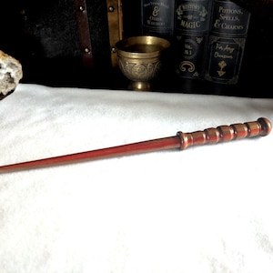 May include: A slender, reddish-brown wooden wand featuring a smooth, tapered shaft and a handle with a series of six rounded, segmented ridges. The wand is positioned horizontally against a white surface, showcasing its polished, multi-toned wood grain.