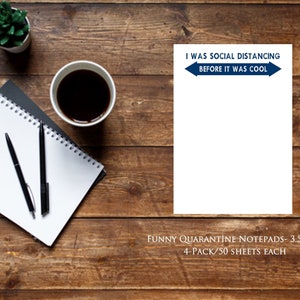 May include: A notepad with the text "I WAS SOCIAL DISTANCING BEFORE IT WAS COOL" in blue. The notepad is 3.5 x 5 inches. The image also shows a cup of coffee, a small plant, two pens, and a spiral notebook on a wooden surface.