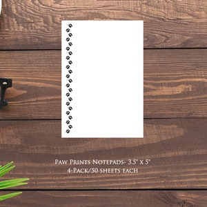 May include: White notepad with black paw print border, 3.5 x 5 inches. The notepad is on a wooden surface, with a cup of coffee, glasses, and a computer mouse visible. The text on the notepad reads "PAW PRINTS NOTEPADS- 3.5" x 5" 4-PACK/50 SHEETS EACH."