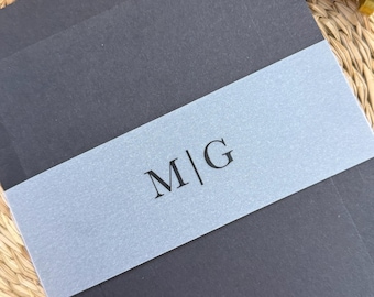 May include: A stack of dark gray envelopes with a light gray band featuring the initials "M|G". A wooden and brass wax seal is visible in the upper right corner. The envelopes are on a woven surface.