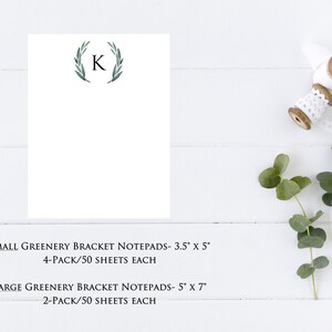 May include: A white notepad with a green leafy wreath and the letter "K" in black. The notepad is on a white wooden surface. The text "SMALL GREENERY BRACKET NOTEPADS- 3.5" X 5" 4-PACK/50 SHEETS EACH" and "LARGE GREENERY BRACKET NOTEPADS- 5" X 7" 2-PACK/50 SHEETS EACH" is printed below the notepad.