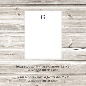 May include: A white notepad with a blue letter "G" printed on the top. The notepad is on a wood background. The text below the notepad reads "SMALL MODERN INITIAL NOTEPADS - 3.5" x 5" 4-PACK/50 SHEETS EACH LARGE MODERN INITIAL NOTEPADS - 5" x 7" 2-PACK/50 SHEETS EACH".