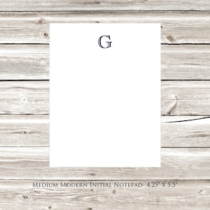 May include: A white notepad with a navy blue letter "G" printed on the front. The notepad is on a light wood background. The text "MEDIUM MODERN INITIAL NOTEPAD - 4.25" x 5.5" is printed below the notepad.