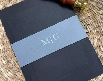 May include: Black stationery set with a light gray belly band printed with the initials "M|G". A wooden handled wax stamp and a wax seal with the same initials are also present. The items are arranged on a woven surface.