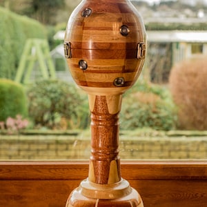 Op de afbeelding: Een decoratief houten object met een ronde bovenkant en een hoge, cilindrische basis. De bovenkant is gemaakt van meerdere stukken hout, die een geometrisch patroon creëren. De basis is ook gemaakt van meerdere stukken hout, die een gestreept patroon creëren. Het object is een uniek en handgemaakt kunstwerk.