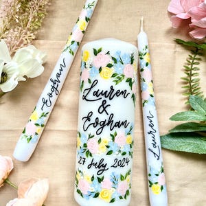 May include: Three white candles, including two tapers and a pillar candle, adorned with floral patterns in pink, yellow, and blue. The pillar candle is inscribed with the names "Lauren & Eoghan" and the date "27 July 2024".