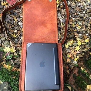 May include: A brown leather messenger bag with a flap closure and a shoulder strap. The bag is open and a black iPad is inside.