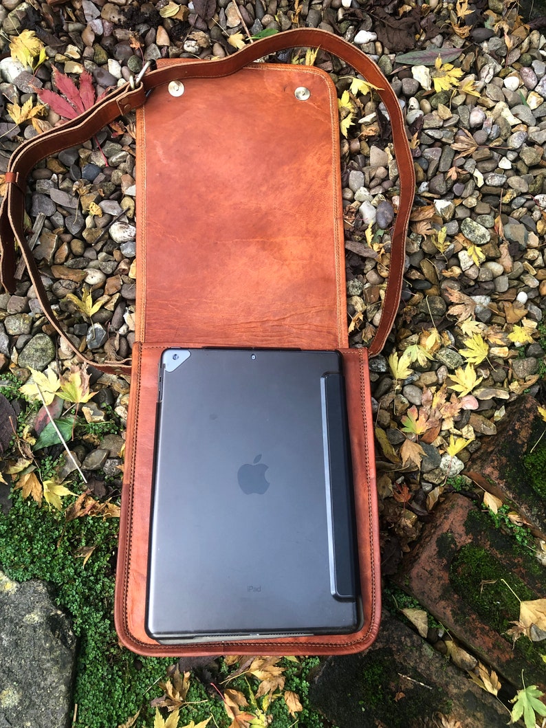 May include: A brown leather messenger bag with a flap closure and a shoulder strap. The bag is open and a black iPad is inside.