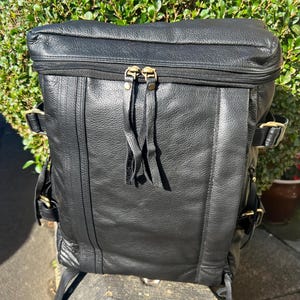 May include: Black leather backpack with a zip closure and adjustable straps. The backpack features brass-coloured hardware and decorative tassels on the zip pulls. The bag is made of textured leather.