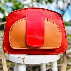 May include: A red and tan leather shoulder bag with a flap closure. The bag has a rounded shape with a tan front pocket and a red flap. The strap is a light tan colour. The bag is sitting on a white stool.