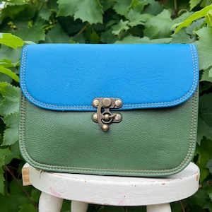 May include: A rectangular crossbody bag featuring a bright blue top flap and a muted olive green body. The bag is constructed with a textured leather-like material and is secured by a central antique brass-toned metal flip-lock clasp closure.