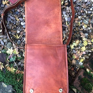 May include: A brown leather messenger bag with a long adjustable strap. The bag has a flap closure and two silver metal snaps on the front.