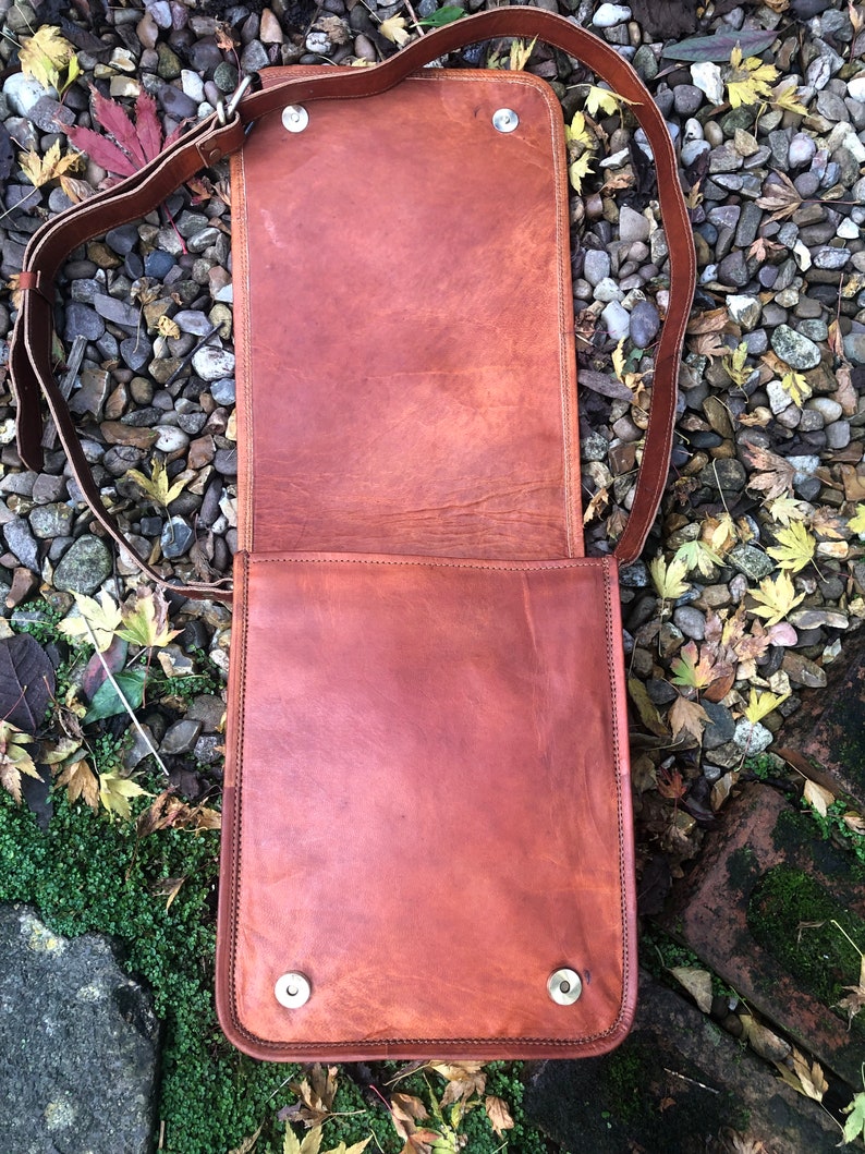 May include: A brown leather messenger bag with a long adjustable strap. The bag has a flap closure and two silver metal snaps on the front.