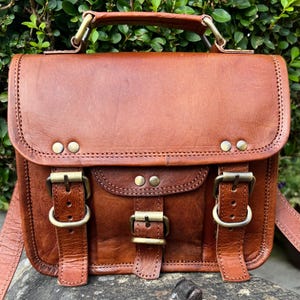 May include: A brown leather satchel featuring a top handle, a fold-over flap with two buckle closures, and a smaller central pocket with a single buckle. The bag is accented with brass-toned metal rivets and hardware throughout the exterior.