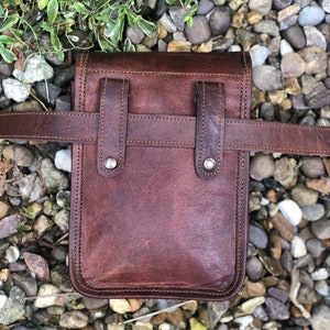 May include: A brown leather fanny pack with a belt loop and two adjustable straps. The bag has a flap with a buckle closure and a small pocket on the front.