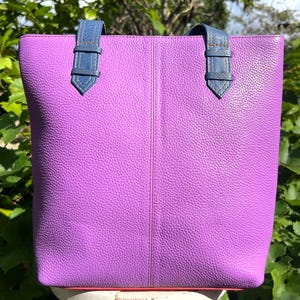 Könnte beinhalten: Eine leuchtend lila Tragetasche mit blauen Ledergriffen. Die Tasche hat eine strukturierte Oberfläche und einen kontrastierenden rosa Besatz am Boden. Die Tasche steht auf einem weißen Hocker vor einem Hintergrund aus grünem Laub.