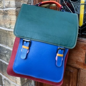 May include: A multi-coloured satchel backpack featuring a dark green top flap, a bright blue main body, and deep red side panels. The bag includes a top handle with orange and purple accents, and two front straps with silver buckles and yellow loops.