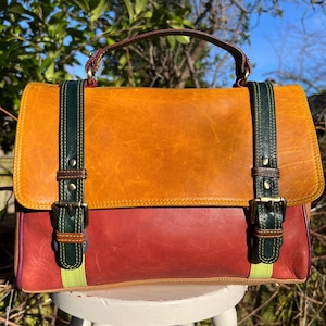 May include: A colorful leather satchel with a top handle and two buckle straps. The bag features a combination of orange, red, and green leather panels. The bag is sitting on a white stool.