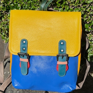 Op de afbeelding: Een rugzak met kleurvlakken, bestaande uit een mosterdgele bovenflap, een felblauwe romp en een geel zijpaneel. De tas heeft twee verticale blauwgroene banden met donkere metalen gespen, afgewerkt met kleine roze en rode lederen details.