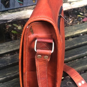 May include: A brown leather messenger bag with a long adjustable strap. The bag has a flap closure and a zippered pocket on the front. The strap has a metal buckle and two metal rivets.