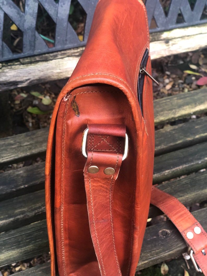 May include: A brown leather messenger bag with a long adjustable strap. The bag has a flap closure and a zippered pocket on the front. The strap has a metal buckle and two metal rivets.