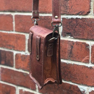 May include: A brown leather crossbody bag with a flap closure and a long adjustable strap. The bag has a small pocket on the front and a buckle closure.