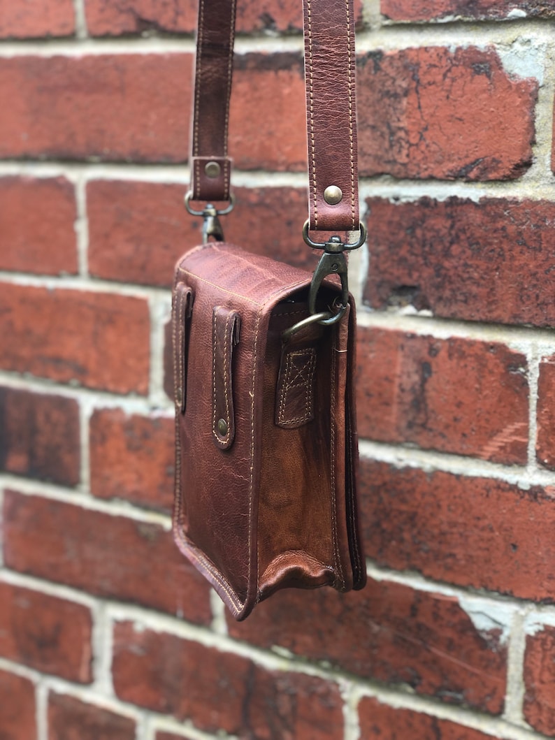 May include: A brown leather crossbody bag with a flap closure and a long adjustable strap. The bag has a small pocket on the front and a buckle closure.