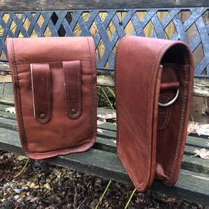May include: Two brown leather bags with a flap closure and a strap. The bag on the left has two pockets on the front. The bag on the right has a metal ring on the strap.