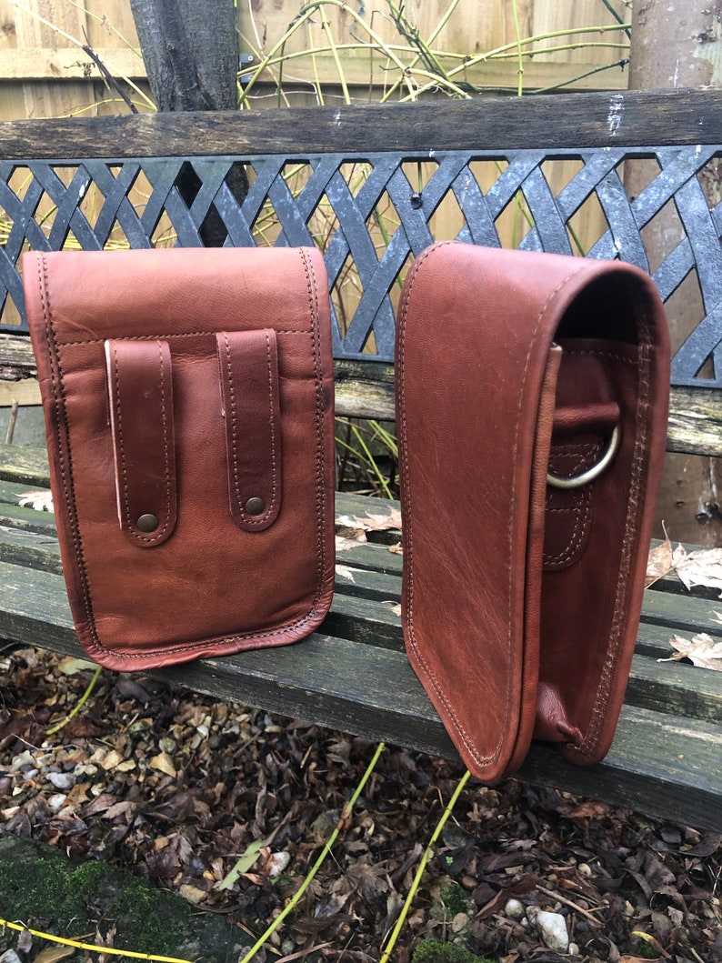 May include: Two brown leather bags with a flap closure and a strap. The bag on the left has two pockets on the front. The bag on the right has a metal ring on the strap.