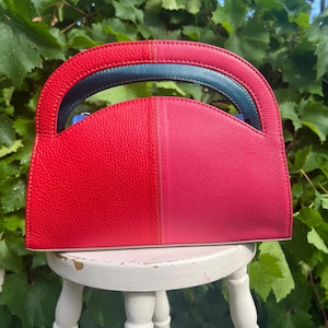 May include: A semi-circular handbag featuring a textured leather exterior with a colour-blocked design in vibrant red and pink. The bag includes a dark blue integrated handle and a flat base, showcasing a distinct two-tone aesthetic throughout.