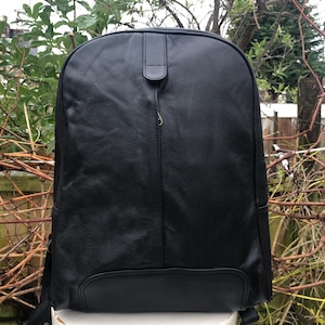 Könnte beinhalten: Ein schwarzer Lederrucksack mit abgerundeter Oberseite und kleinem Klappenverschluss. Der Rucksack hat eine vertikale Naht in der Mitte und eine kleine Tasche unten. Die Träger sind an den Seiten sichtbar. Die Tasche ist aus strukturiertem Leder.