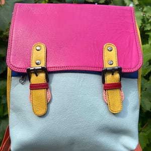 May include: A multi-colored backpack featuring a bright pink top flap, a light blue main body, and yellow side panels. It includes two yellow leather straps with metal buckles, red accents, a green top handle, and orange shoulder straps.
