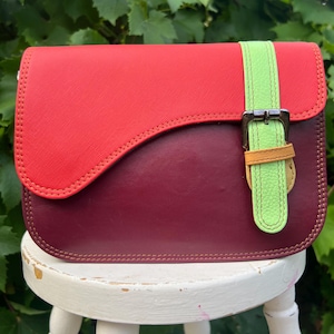 May include: A structured crossbody bag featuring a vibrant red flap with a textured finish over a smooth deep burgundy body. A bright lime green vertical strap with a silver metal buckle and a small tan leather loop secures the front closure.