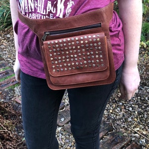 May include: A brown leather belt bag with a zippered pocket and a front flap covered in silver studs. The bag is worn around the waist of a person wearing a purple shirt and black jeans.
