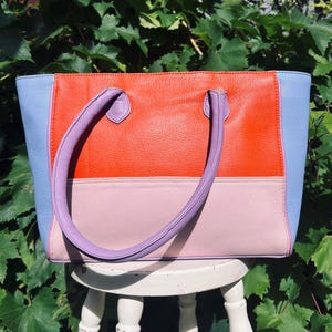 May include: A colour-blocked tote bag featuring a vibrant orange upper section, a soft pink lower section, and light blue side panels. The bag includes two lavender handles attached to the top, with a textured surface throughout the exterior.