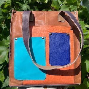 May include: A rectangular leather tote bag featuring a brown base with a large orange panel and two exterior pockets in bright turquoise and royal blue. The bag includes two dark purple shoulder straps and silver-toned metal rivets on the pockets.