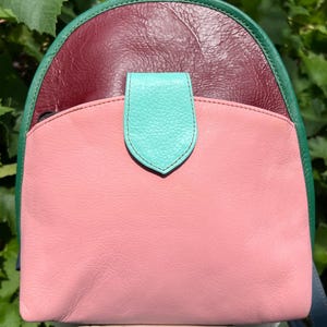 Op de afbeelding: Een kleine leren rugzak met een color-block ontwerp, bestaande uit een donkerbordeaux bovendeel, een lichtroze voorvak en een fel turquoise sluiting. De tas is afgewerkt met een donkergroene leren rand en is voorzien van een zwarte ritssluiting.
