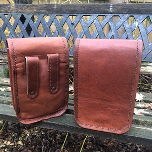 May include: Two brown leather bags with a flap closure. The bag on the left has two pockets on the front. Both bags have a stitched edge.