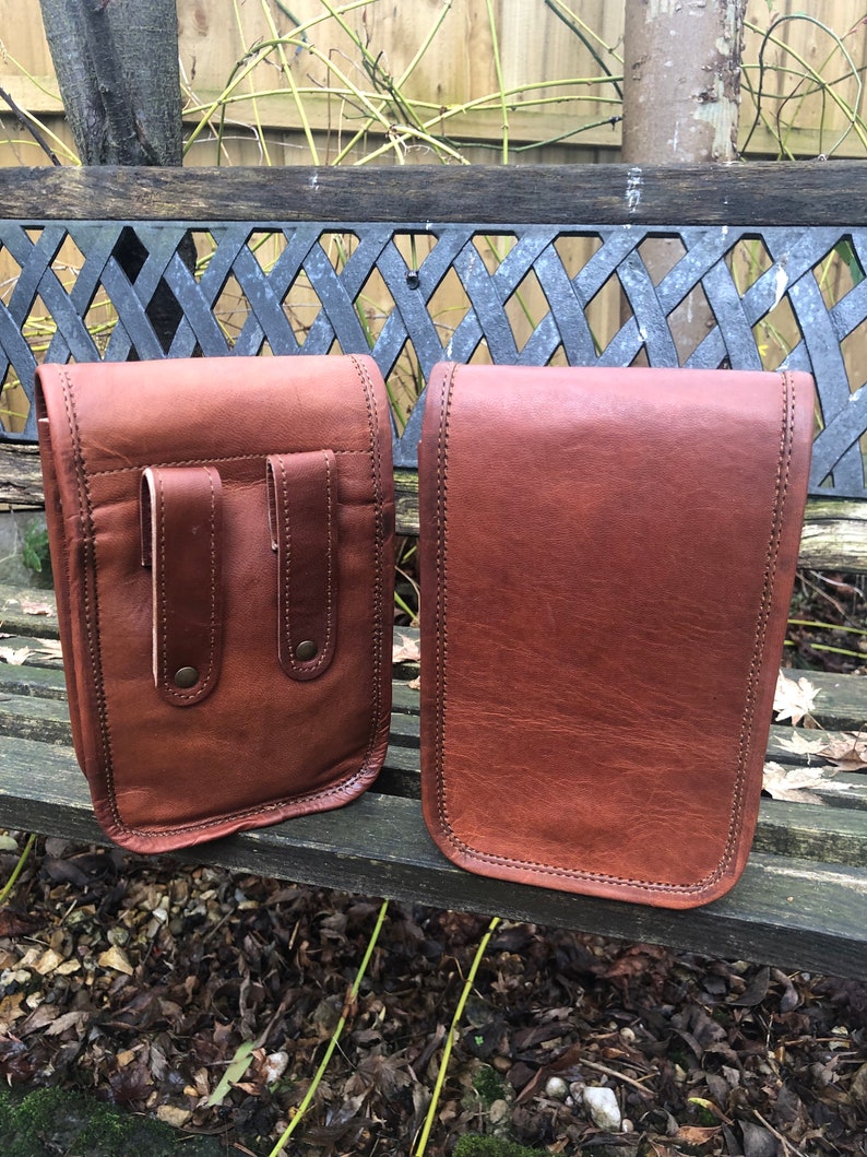 May include: Two brown leather bags with a flap closure. The bag on the left has two pockets on the front. Both bags have a stitched edge.