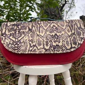 May include: A red and snakeskin-patterned clutch bag with a teal handle. The bag has a curved shape with a red leather bottom and a snakeskin-patterned flap. The bag is sitting on a white stool.