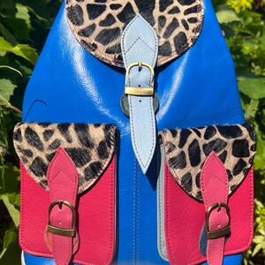 May include: A vibrant blue backpack featuring a top flap and two front pockets with a black and tan animal print pattern. The bag includes light blue and pink leather straps with brass-toned metal buckles, providing a secure closure for each compartment.
