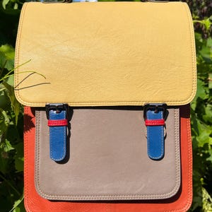 May include: A multi-coloured satchel featuring a large yellow top flap, a central grey pocket, and an orange base. The bag includes a bright pink top handle, two blue vertical straps with red accents, and dark metal buckles for closure.