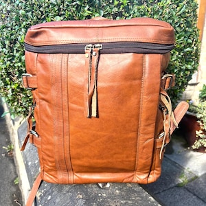 May include: A brown leather backpack with a zipped top and multiple buckle fastenings. The bag has a rectangular shape with a rounded top and features vertical stitching details. The backpack has adjustable straps and a handle.