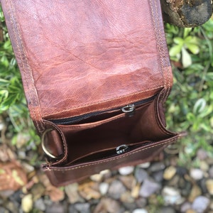 May include: A brown leather crossbody bag with a zippered closure and a metal ring on the side. The bag is open and the inside is visible.