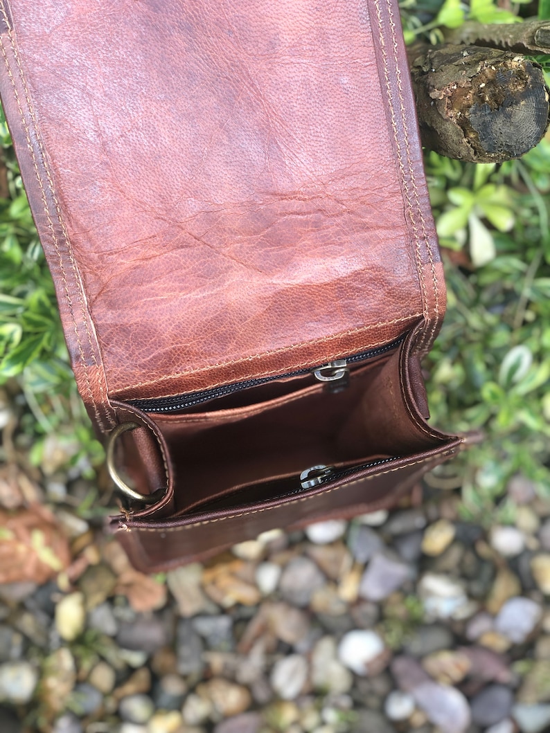 May include: A brown leather crossbody bag with a zippered closure and a metal ring on the side. The bag is open and the inside is visible.