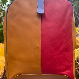 Könnte beinhalten: Zweifarbiges Leder-Rucksack mit Farbblock-Design. Die linke Seite ist hellbraun, die rechte Seite ist leuchtend rot. Der Rucksack hat eine blaue Umrandung, einen lavendelfarbenen Griff und braune Träger. Der Rucksack steht auf einem weißen Hocker.