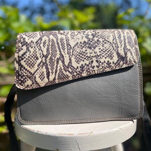 May include: A grey leather crossbody bag with a snakeskin patterned flap in brown and cream. The bag is on a white stool and has a black strap. The background is blurred green.