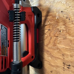 Op de afbeelding: Een rood en zwart elektrisch gereedschap met een zilveren veer en een zwarte slang eraan bevestigd. Het gereedschap is gemonteerd op een houten oppervlak.