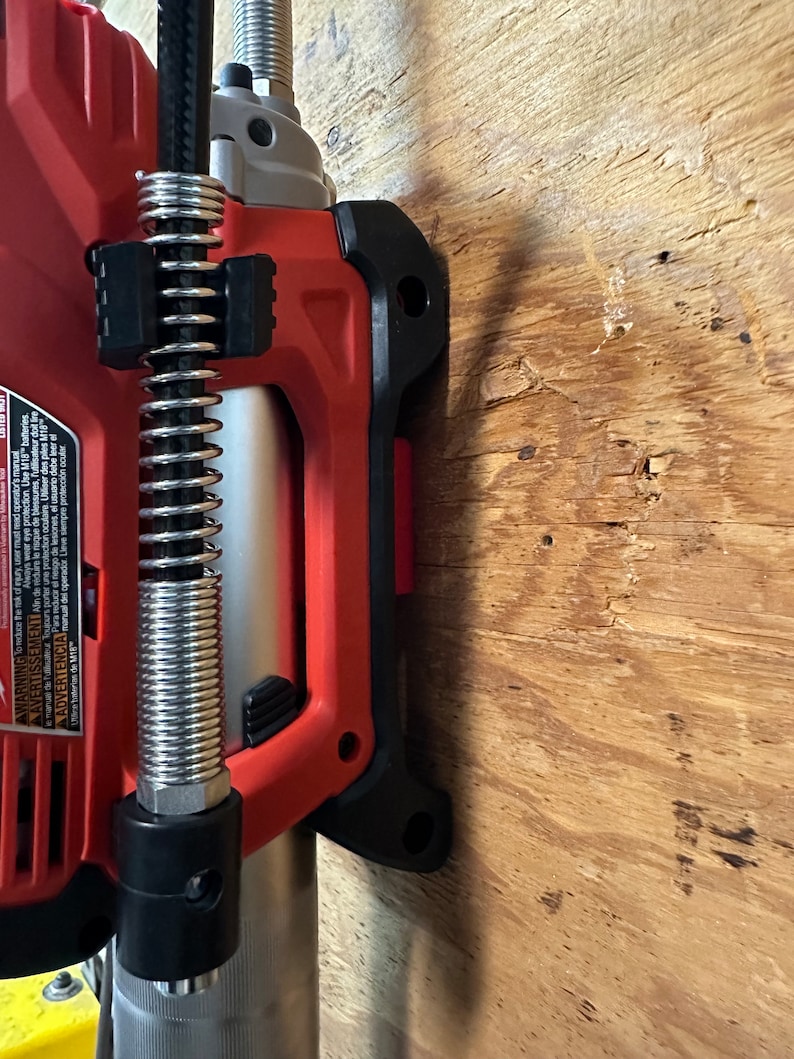 Op de afbeelding: Een rood en zwart elektrisch gereedschap met een zilveren veer en een zwarte slang eraan bevestigd. Het gereedschap is gemonteerd op een houten oppervlak.
